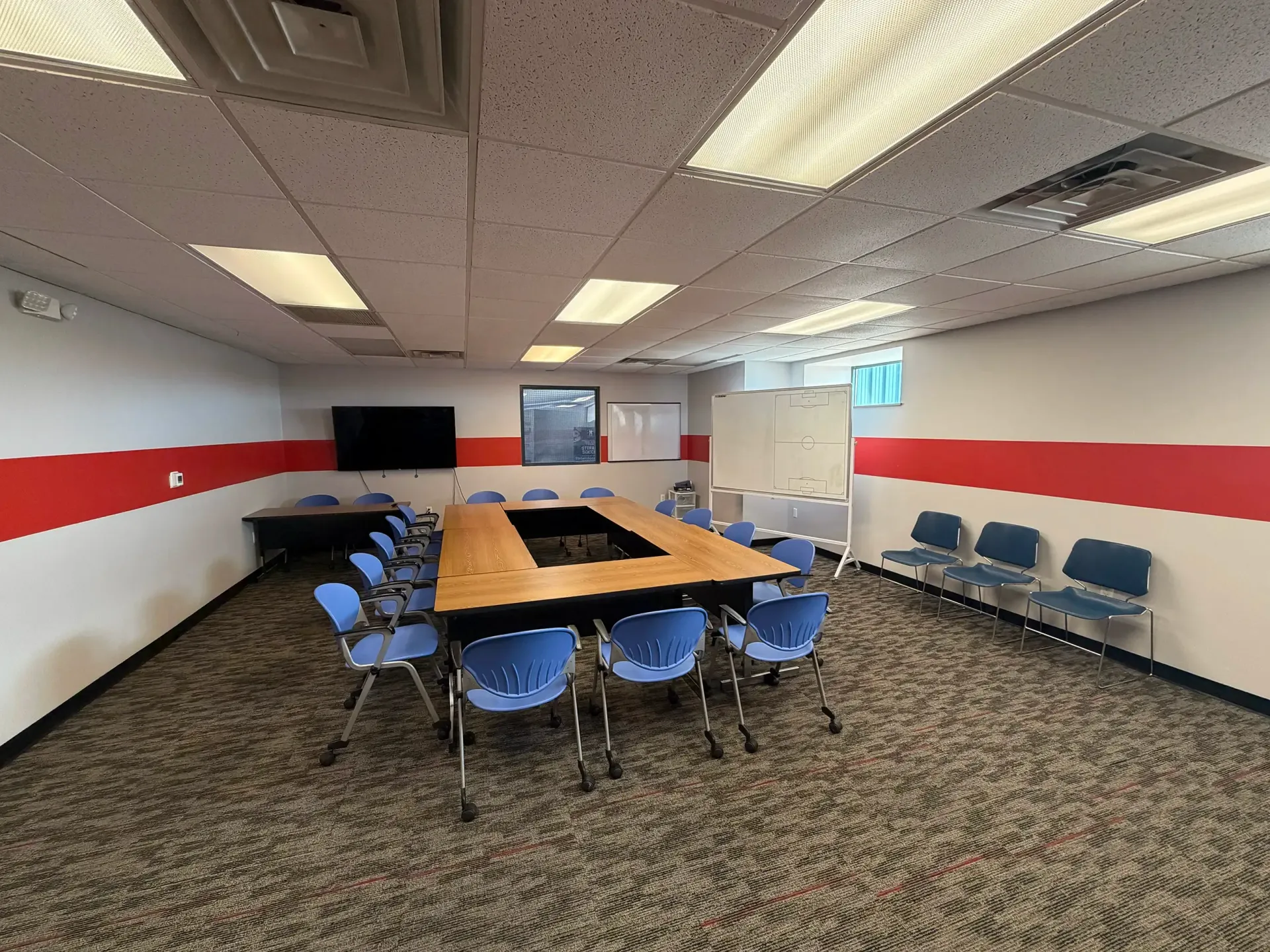 Empty conference room with long table, blue chairs, wall-mounted TV, and red stripe on white walls