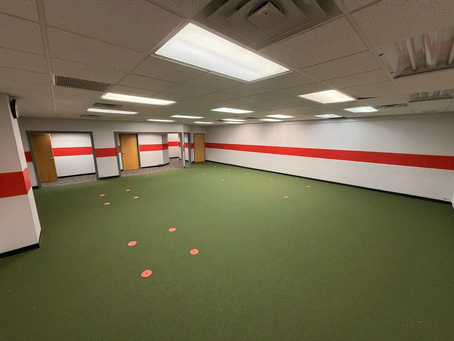 Empty indoor room with green carpet, white walls with red stripe, and ceiling lights