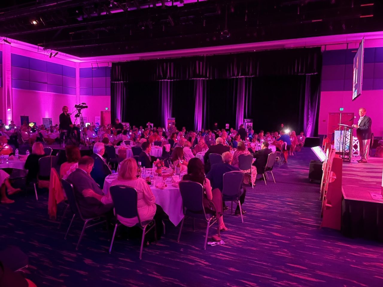 A large group of people are sitting at tables in a large room with purple lights.
