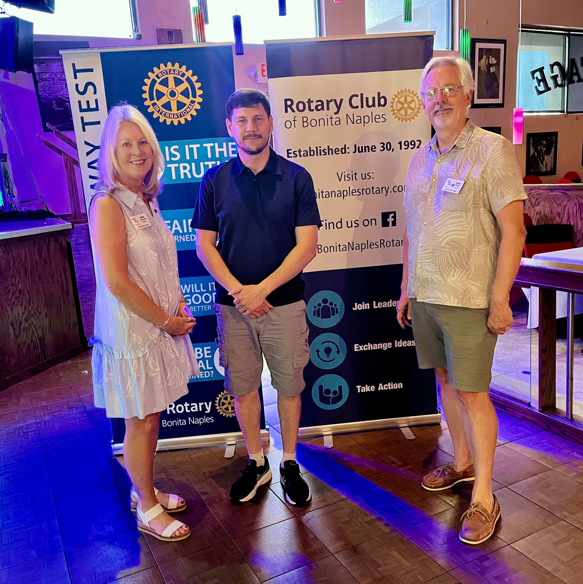 Three people posing at a Rotary Club event in front of a banner and sign