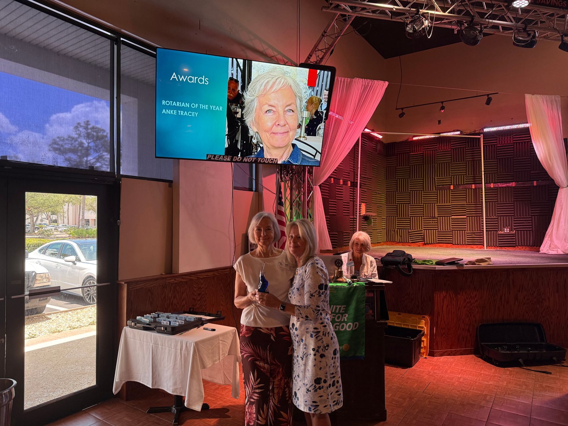 Two people chatting in a dim event hall beside a turquoise screen with a woman's portrait.