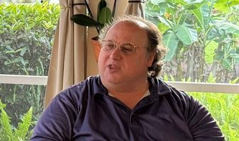 A man wearing glasses and a blue shirt is sitting in front of a window.
