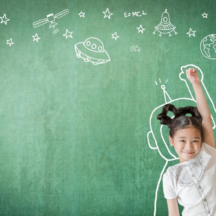 A little girl stands in front of a green chalkboard with drawings on it
