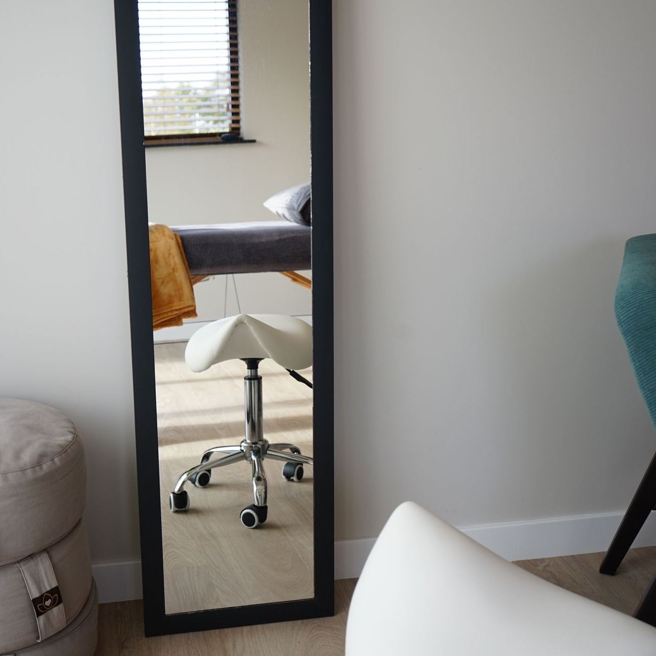 Een grote spiegel met zwart frame weerspiegelt een kamer met een massagetafel en krukje