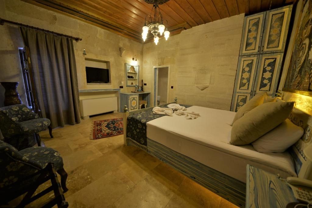 Hotel room with a bed, two armchairs, TV, and decorative panels. Beige walls, wooden ceiling, and patterned rug.