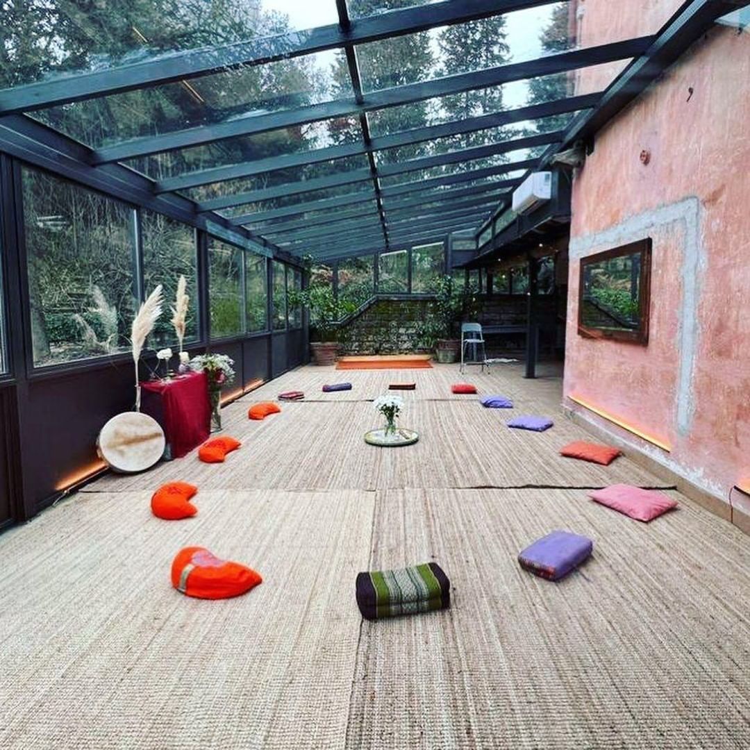 Yoga studio with mats arranged on a textured floor beneath a glass roof, surrounded by plants and a colorful wall.