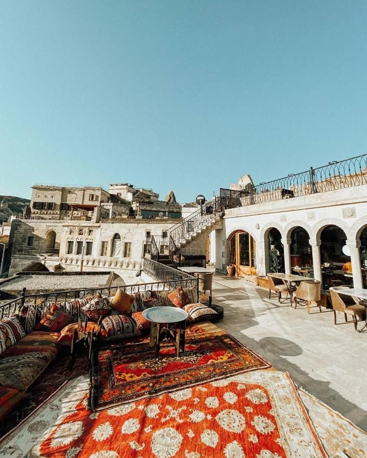 Rooftop lounge with colorful rugs, cushions, and arched windows, overlooking a town with stone buildings under a blue sky.