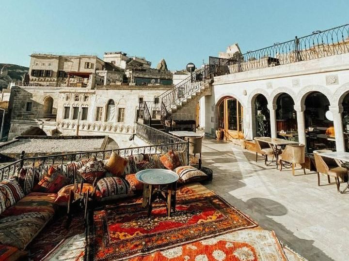 Outdoor terrace with seating, cushions, carpets, and a view of stone buildings in Cappadocia, Turkey.