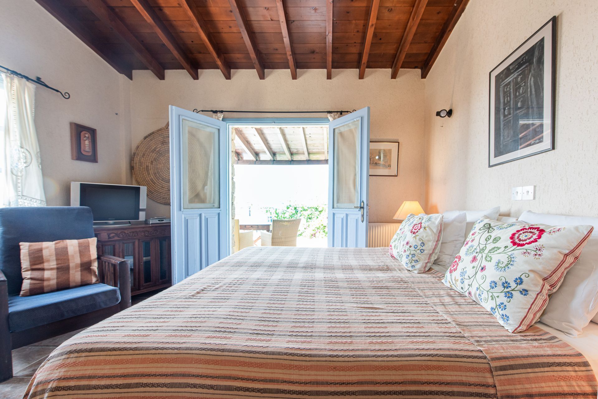 Bedroom with bed, blue doors to a balcony, wooden ceiling, armchair, and artwork.