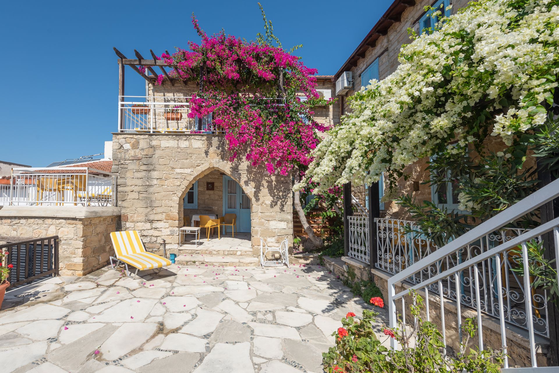 Stone building with patio, purple and white flowering vines, lounge chair, and blue sky.
