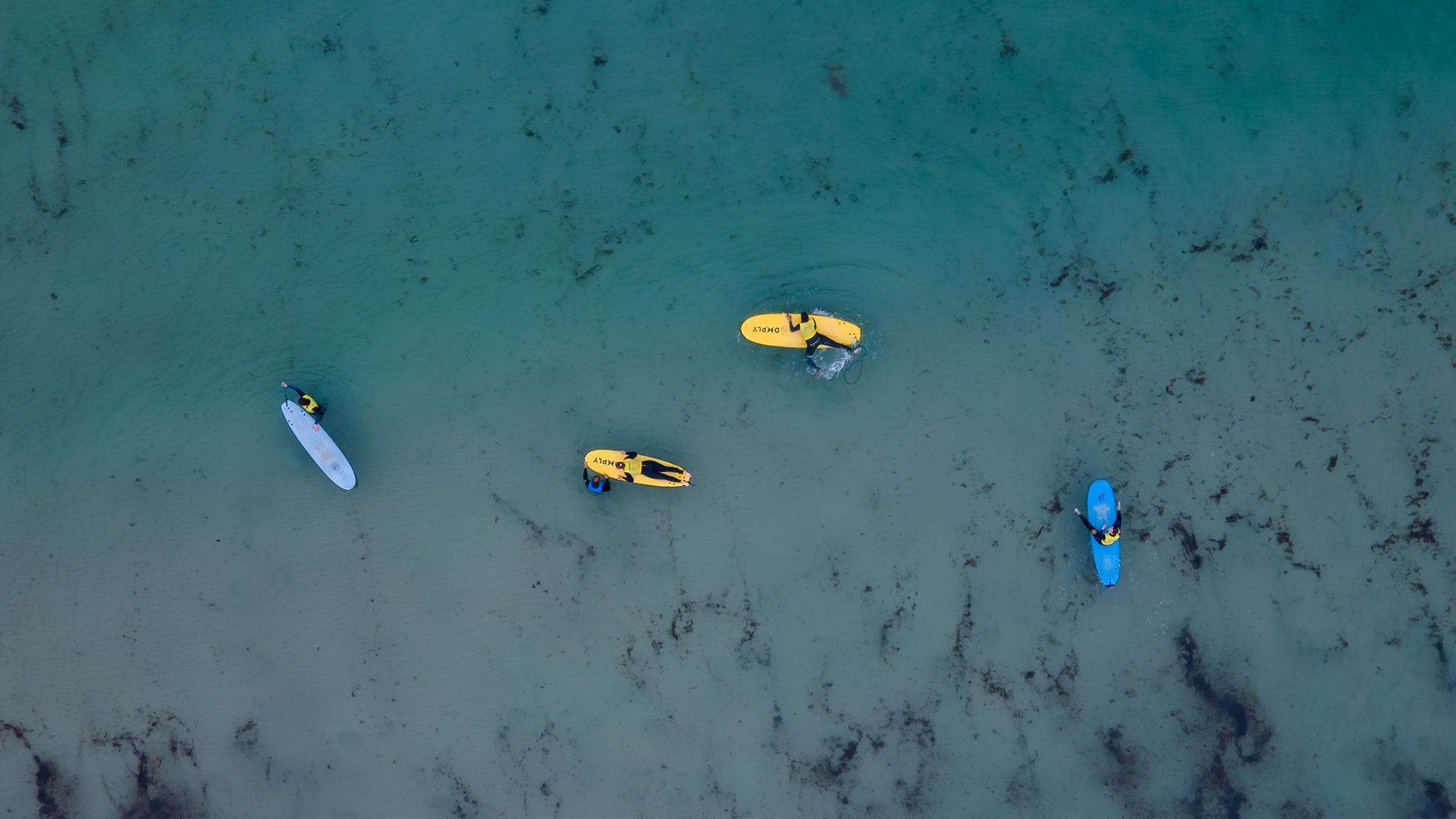Aerial view of shallow turquoise water with a person on a surfboard, two yellow boats, and a blue kayak.