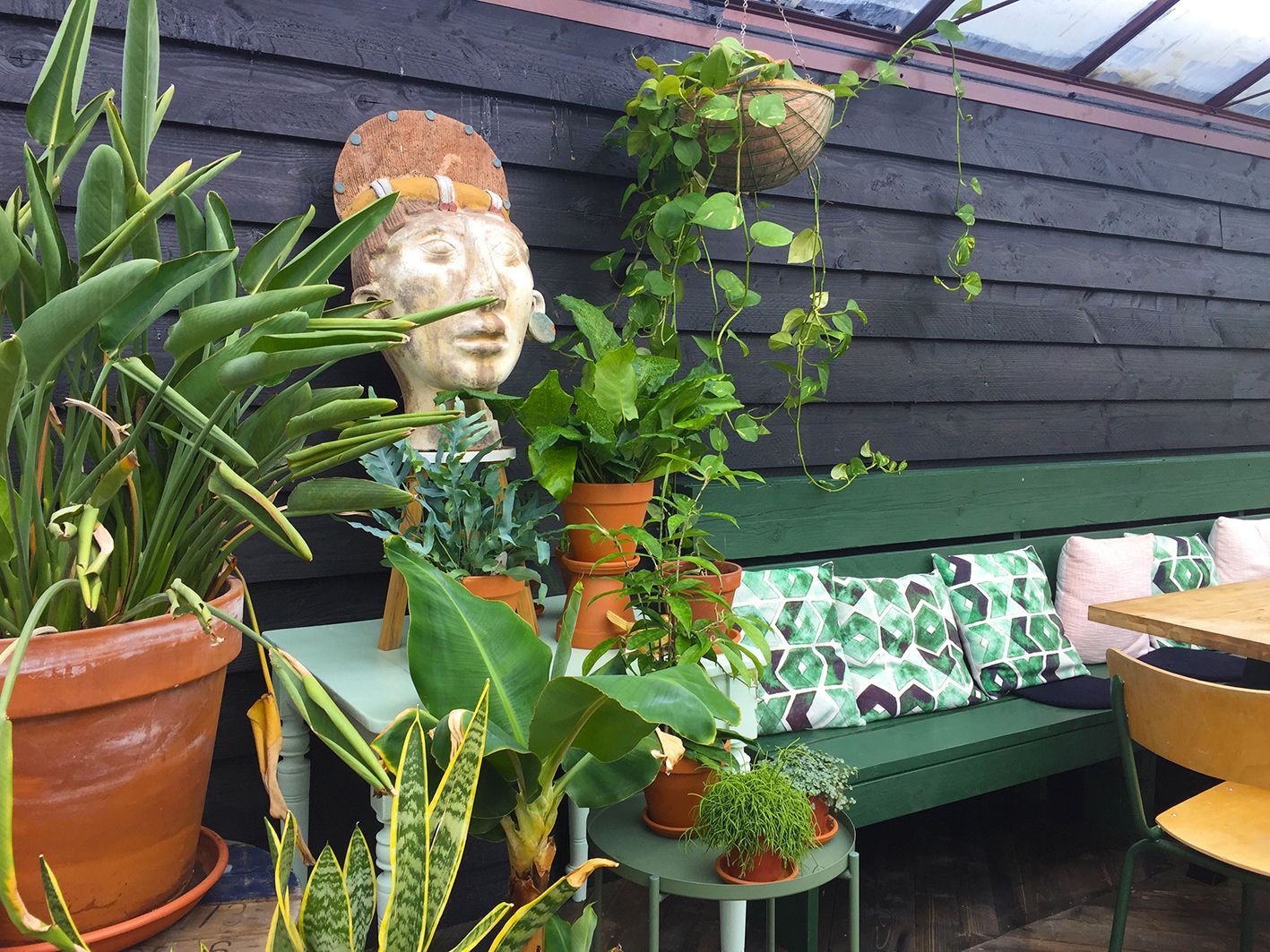 A green bench with pillows, plants in pots, and a sculpture are set against a black wall.