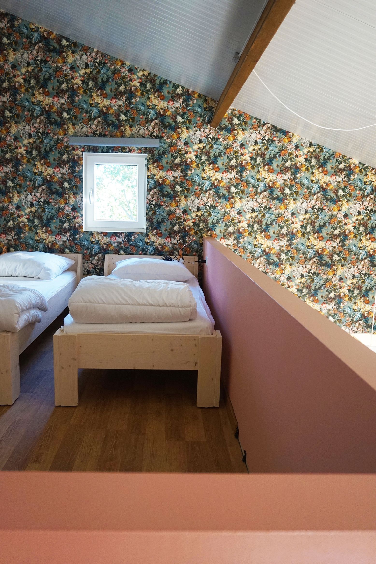 Two beds in a room with floral wallpaper and a small window. Pink railing in foreground.