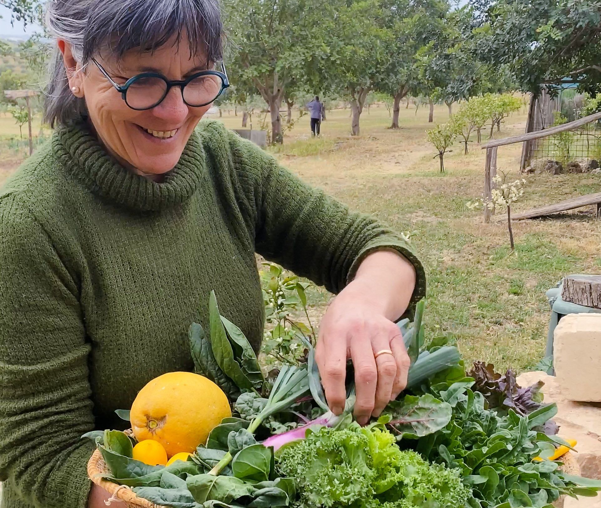 Woman smiles, holding a basket of fresh produce in an orchard.