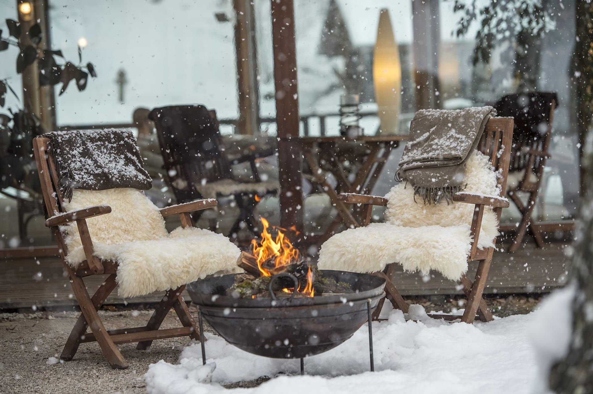 Two wooden chairs with fur throws by a fire pit in snowy setting.