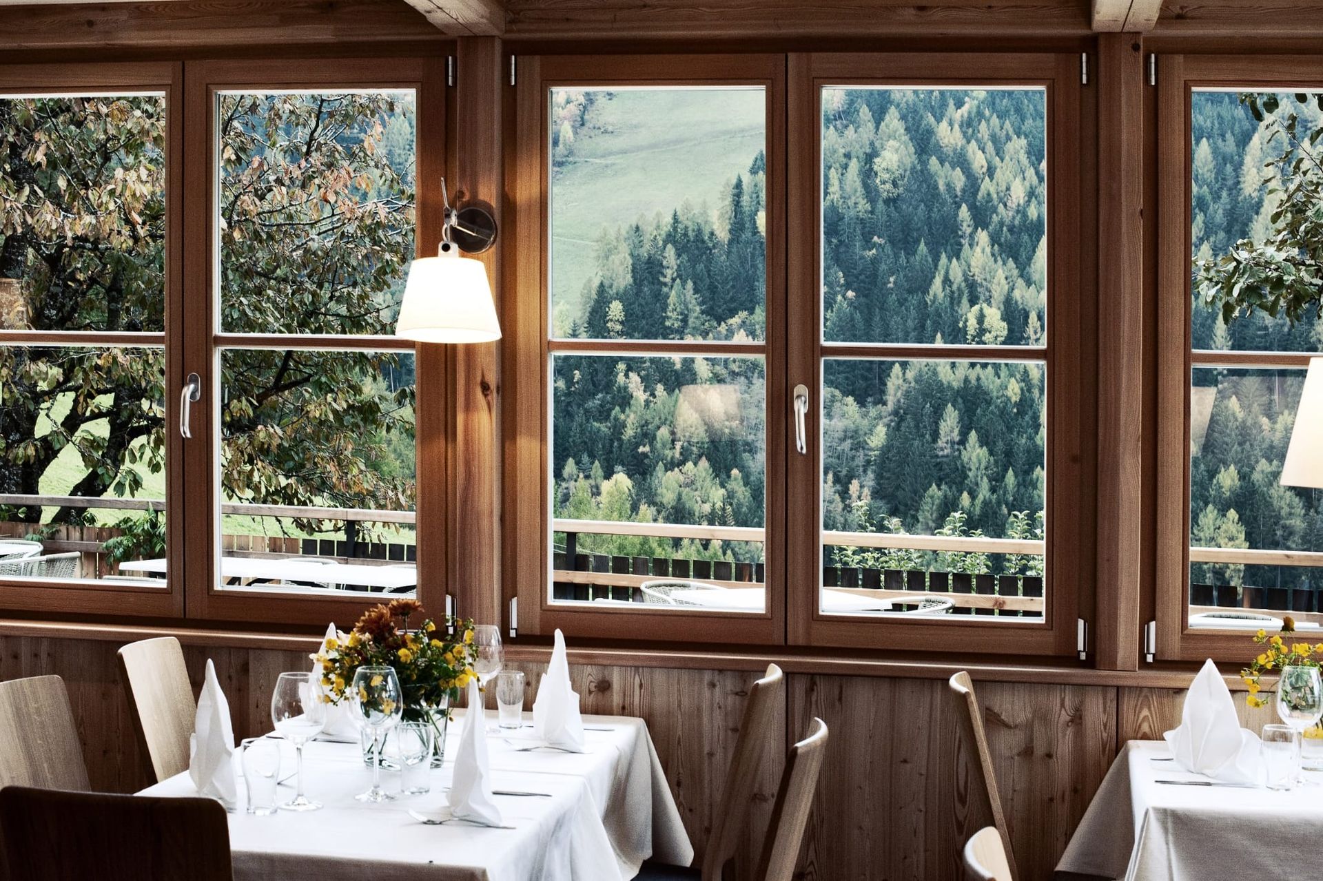 Restaurant interior with wooden frame windows overlooking a green forest and valley; tables set with white linens.