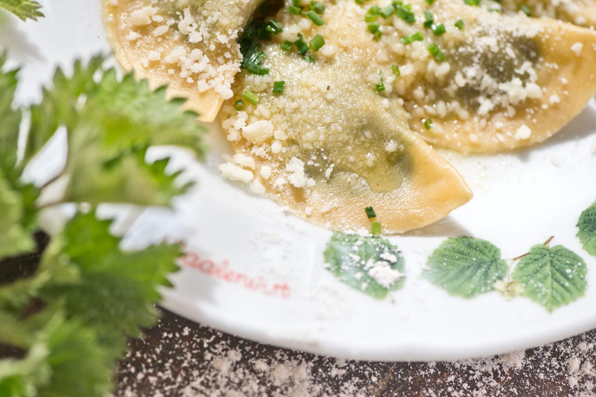 Ravioli with green filling, topped with grated cheese and chives, on a decorated white plate.
