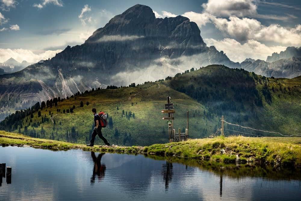 Hiker with backpack walks along a water's edge, mountains in the background, cloudy sky.