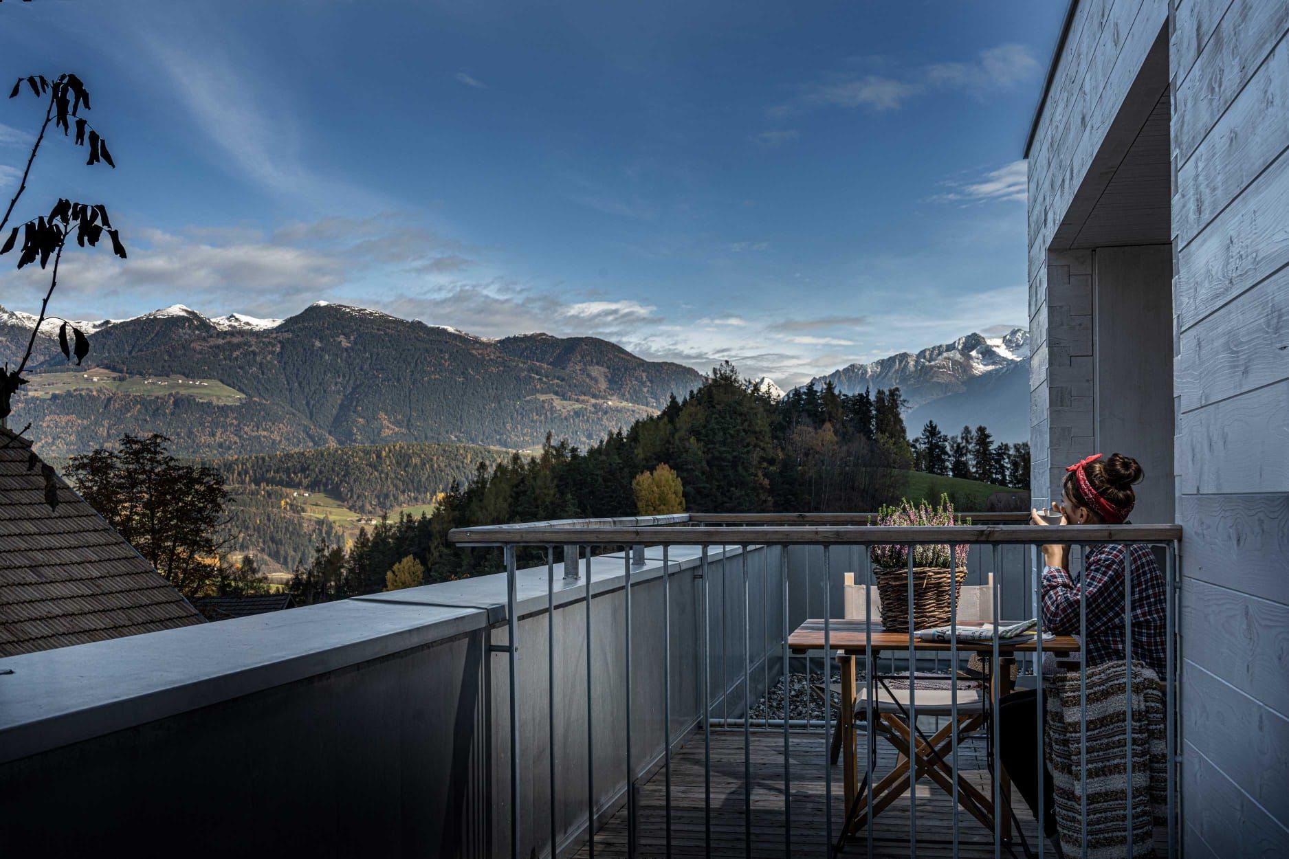 Balcony with table and mountain view under a blue sky, person seated, light tones.