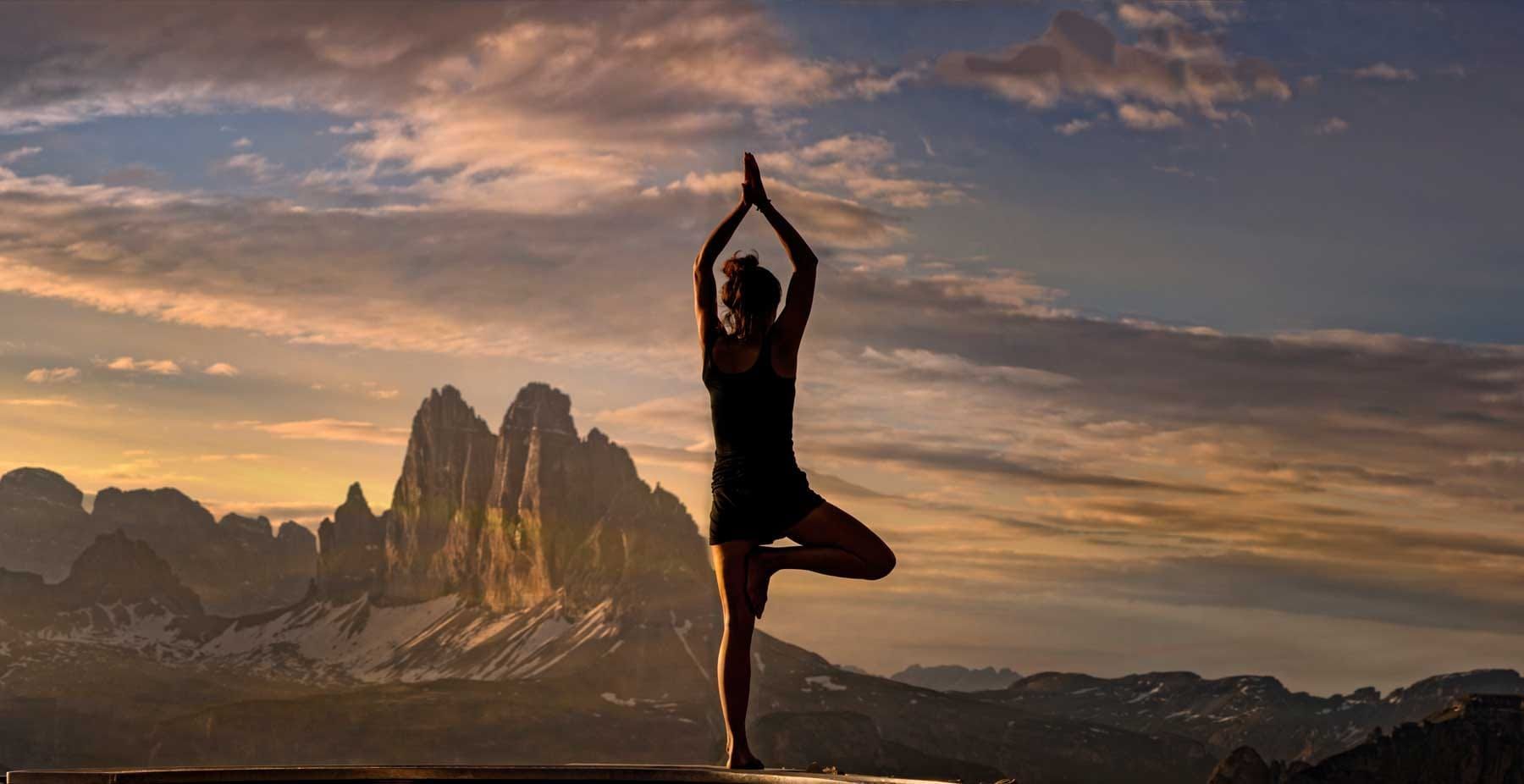 Woman in tree pose, arms raised, with mountains and sunset backdrop.