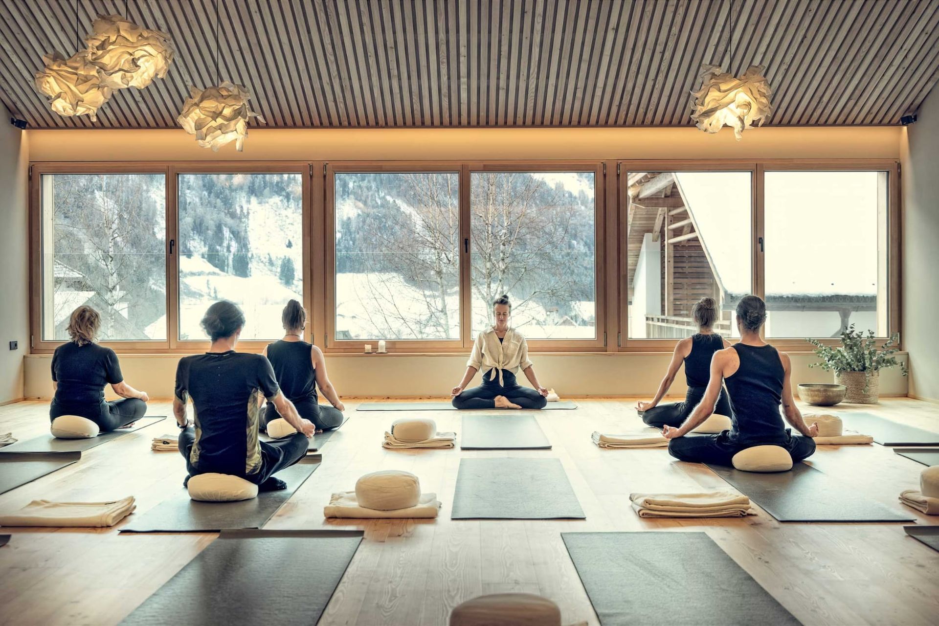 Yoga class in a sunlit room with mountain view. People sit in meditation pose on mats.