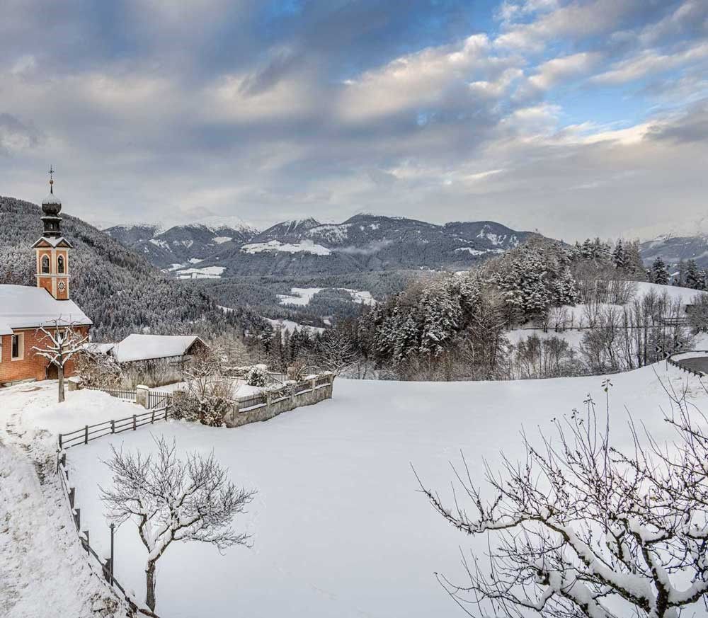 Snow-covered village with church in the foreground and mountains in the background under a cloudy sky.