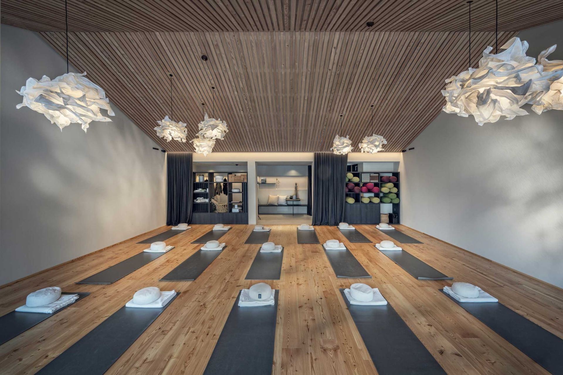 Yoga studio with wooden floor, mats, and overhead cloud-like lights; back wall with shelves and flowers.
