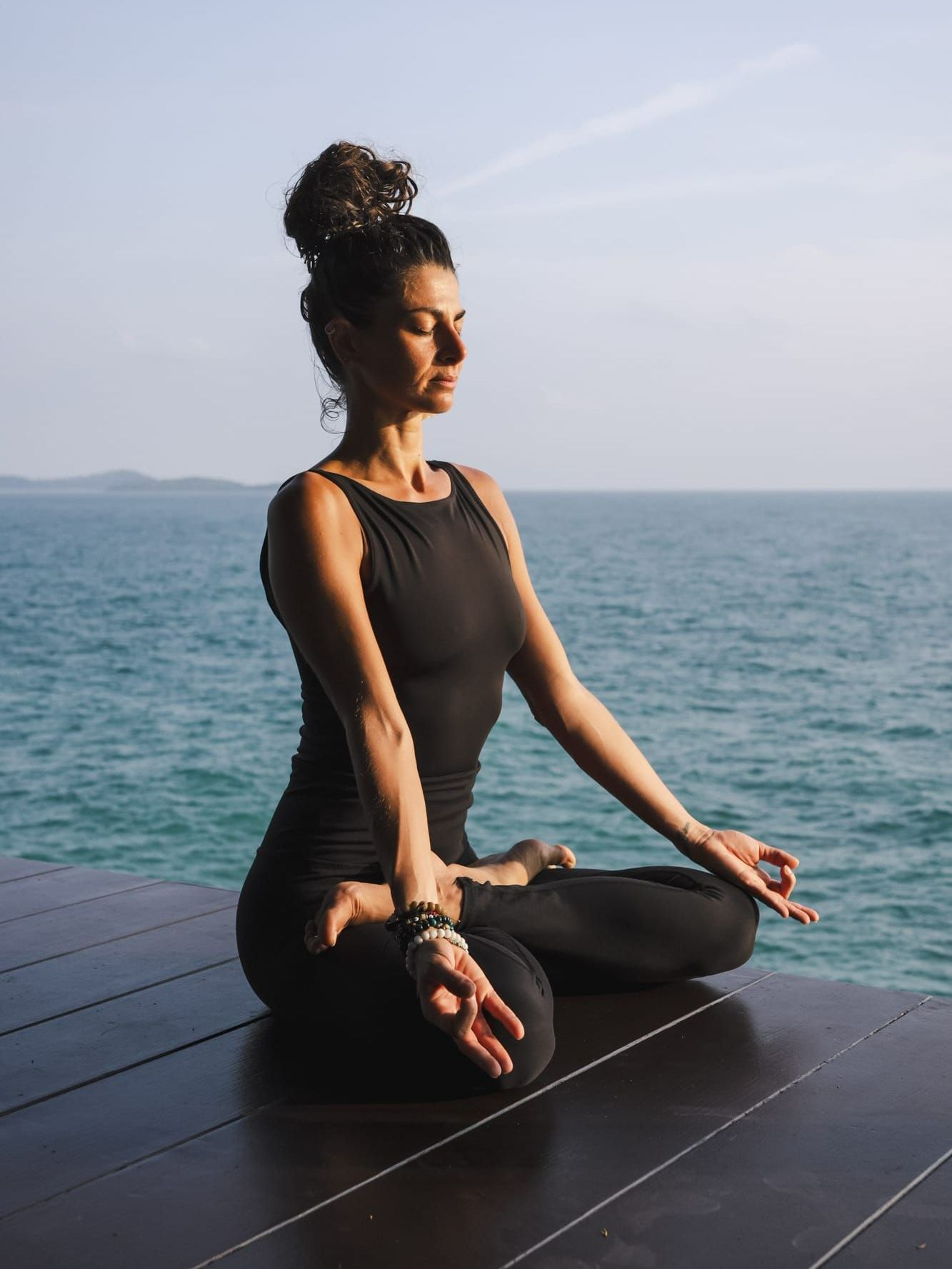 Woman in black yoga attire meditating near ocean, arms extended, eyes closed.
