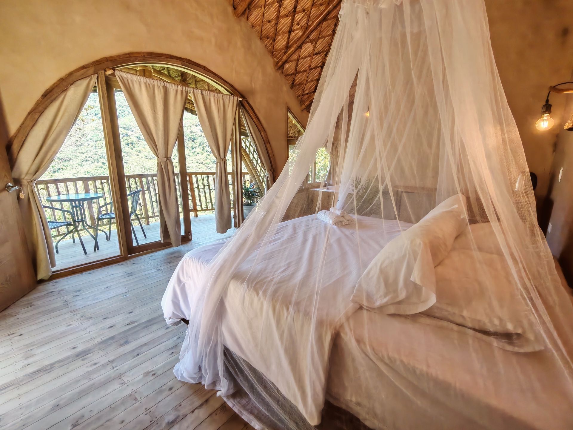 Cozy bedroom with bed under a mosquito net. Arched window overlooks a balcony with a table and chairs.