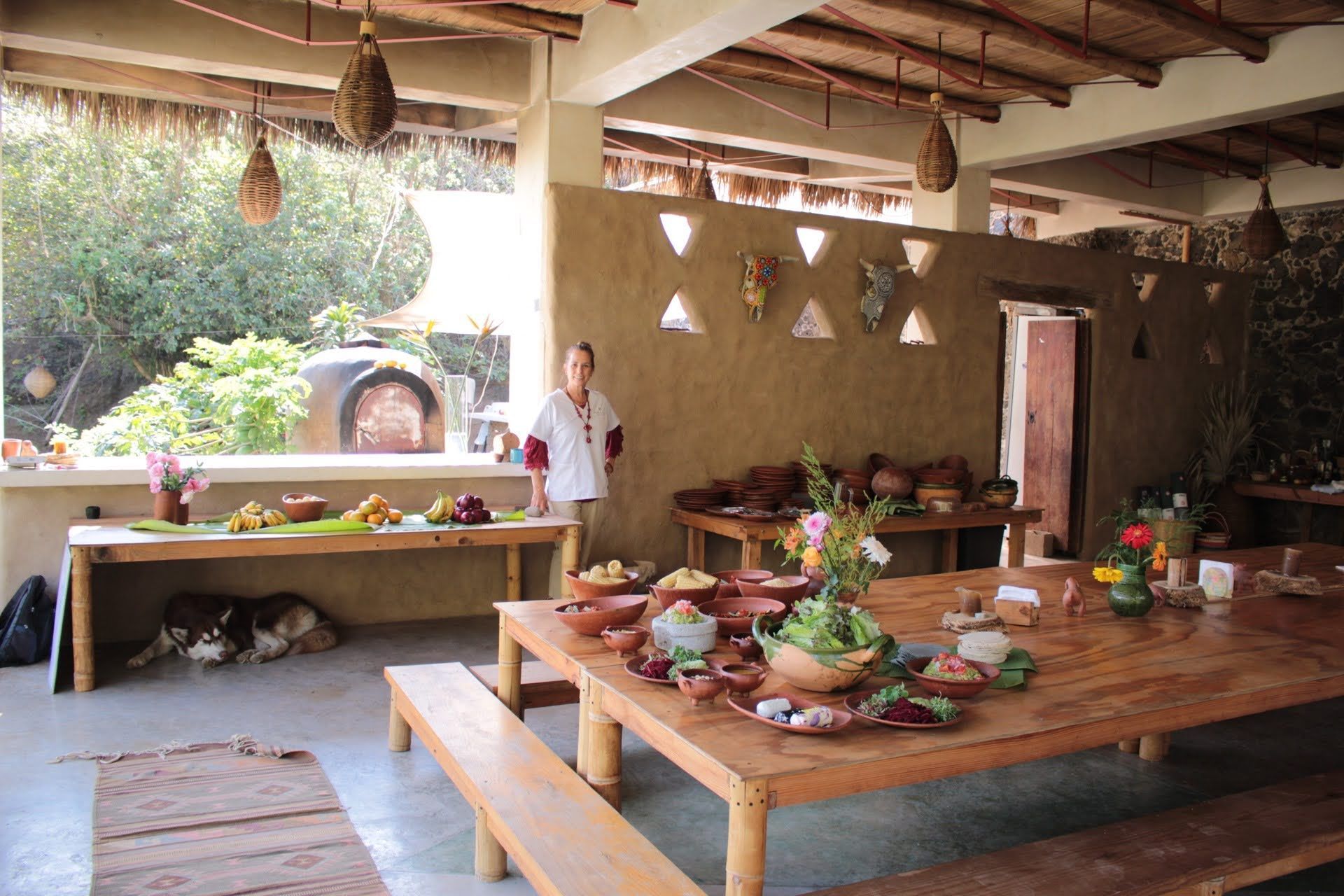Covered outdoor dining area with tables set with food, person standing near a mud wall.