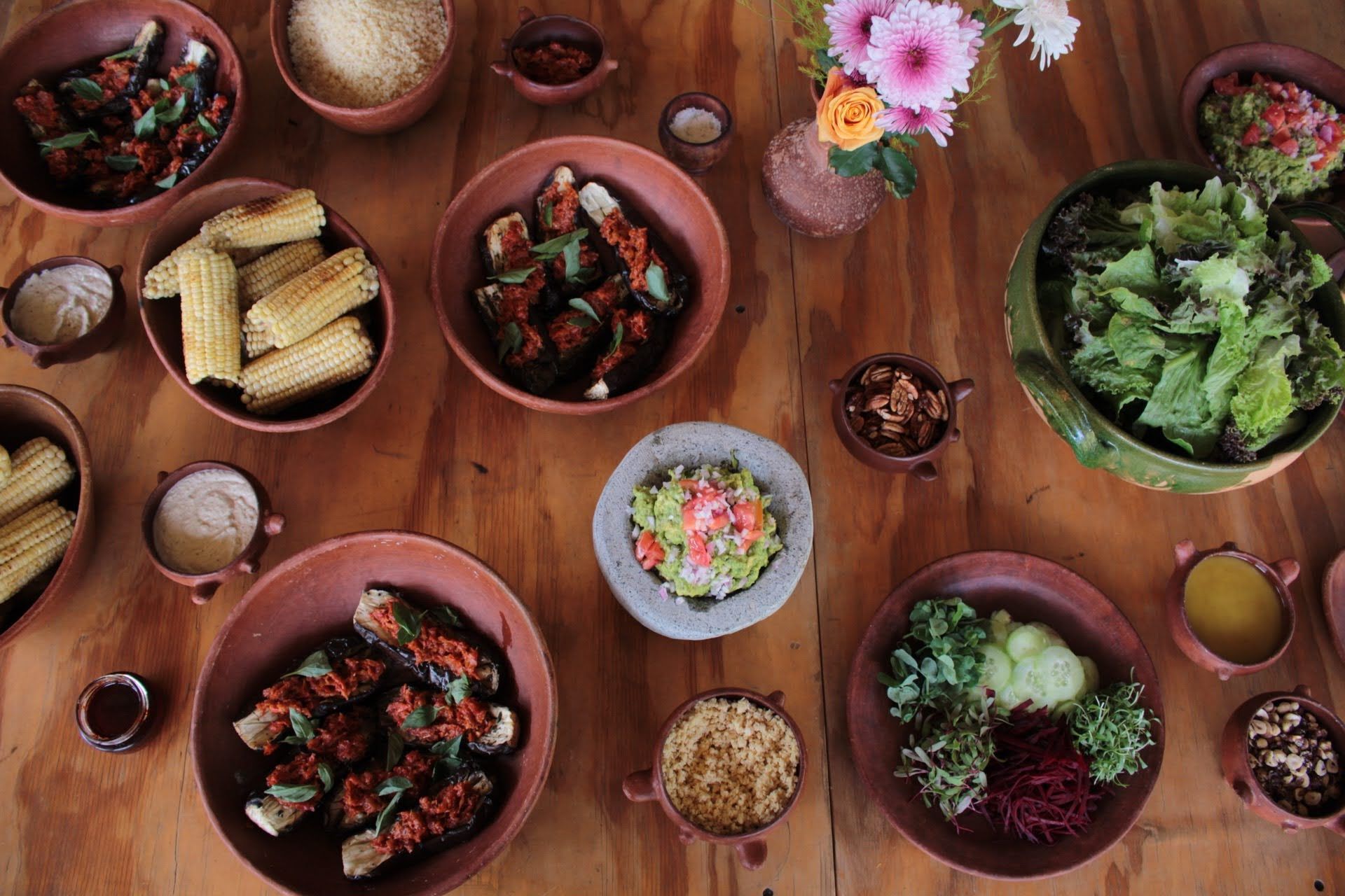 Overhead view of a wooden table laden with various dishes in terracotta bowls, including grilled corn and salads.