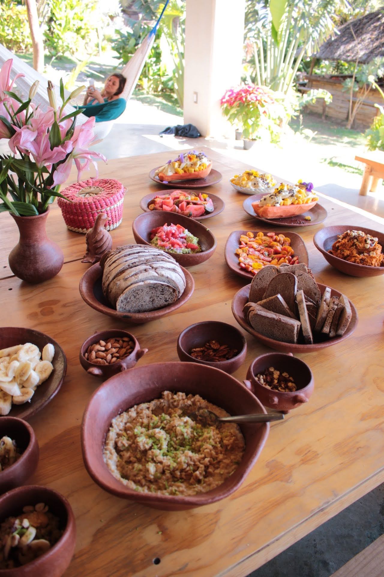Wooden table with spread of food, flowers, and person in hammock in tropical setting.