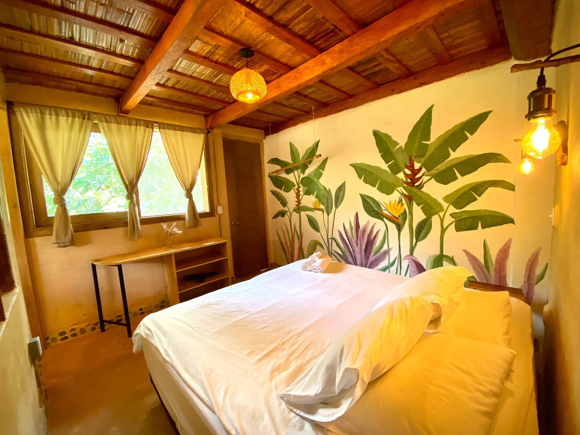 Bedroom with a painted mural of tropical plants above a bed. Windows with curtains and wood ceiling.