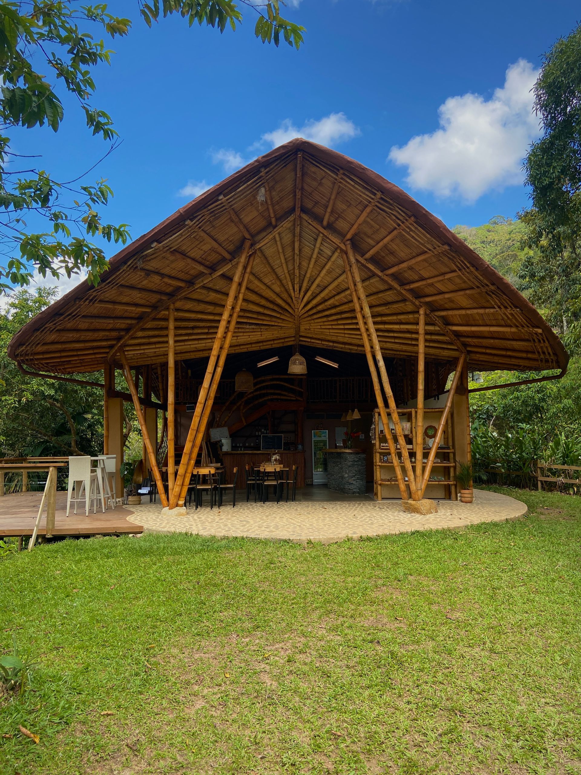 Bamboo-framed open-air structure with a thatched roof, in a grassy area with blue sky in the background.