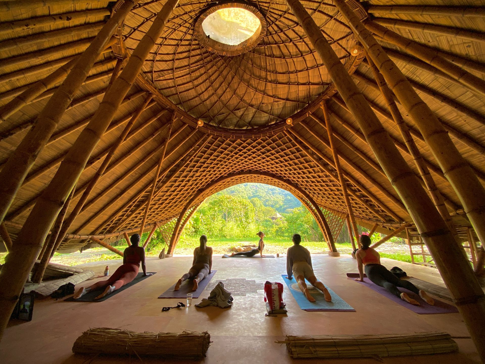 Yoga class in a bamboo structure, practicing upward-facing dog pose. Green foliage visible outside.