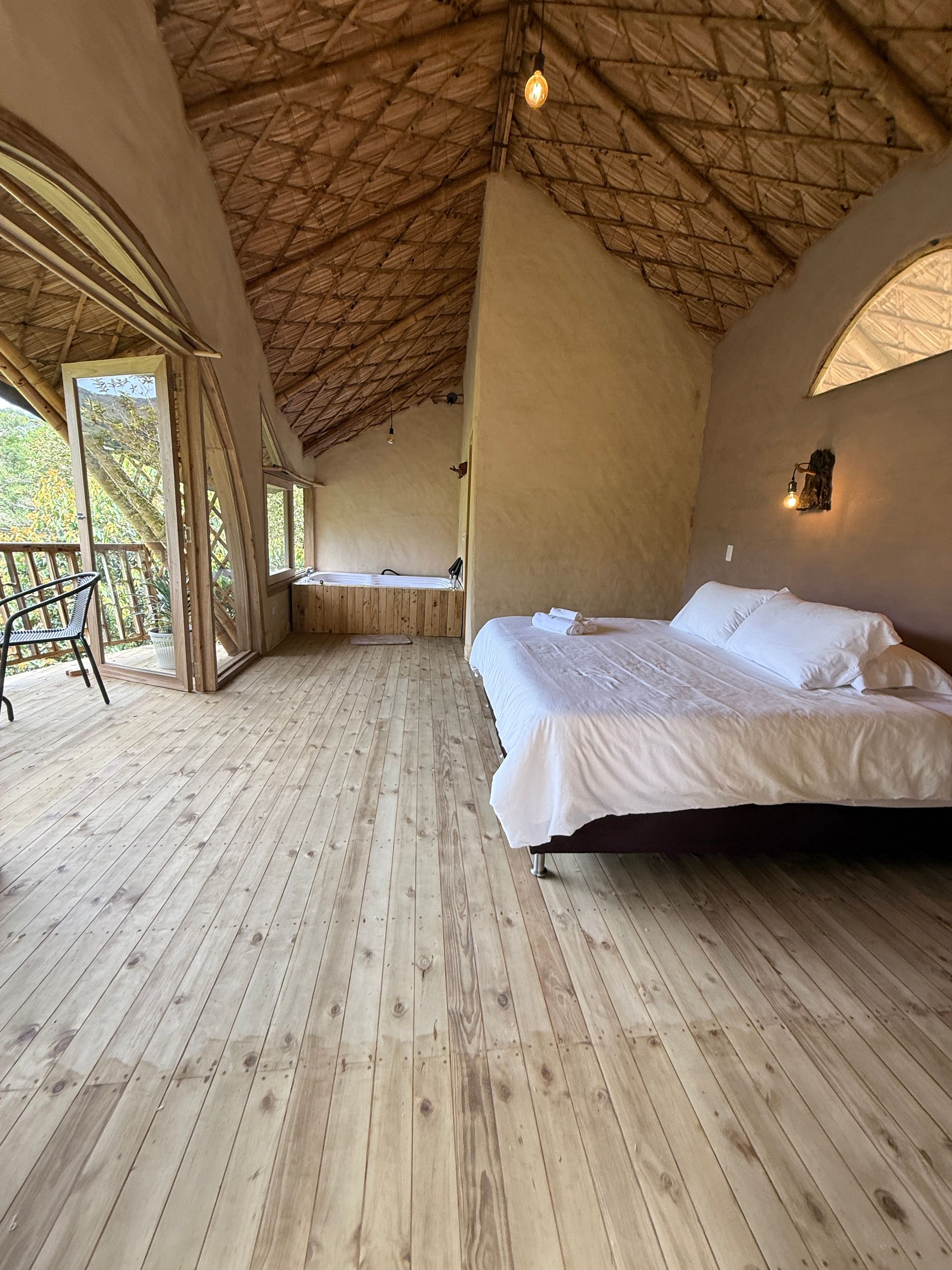 Bedroom with straw roof, wooden floor and bed. Doors open to a balcony overlooking foliage.