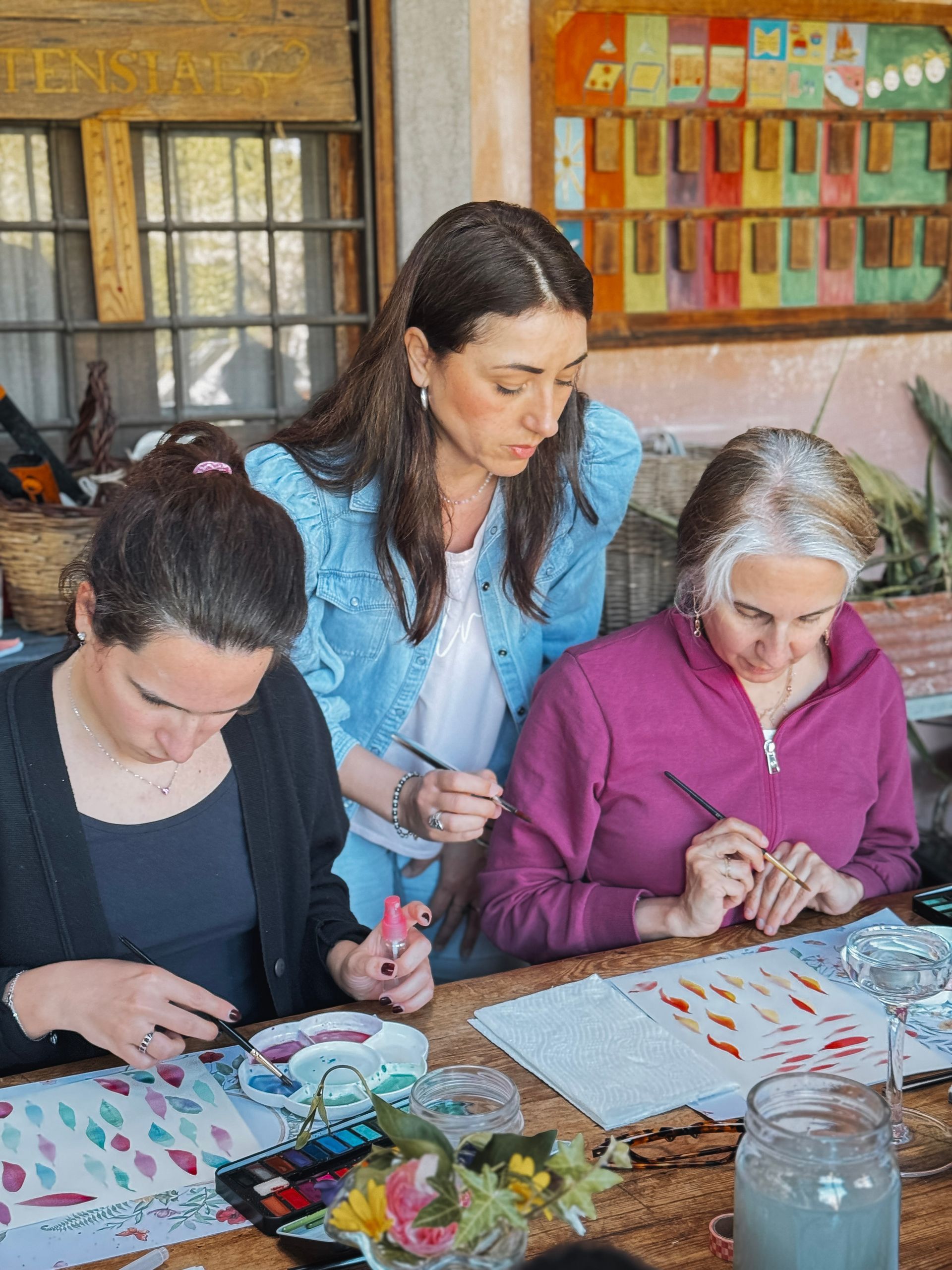 Three people painting at a table outdoors. Woman in blue denim jacket helps other painters.