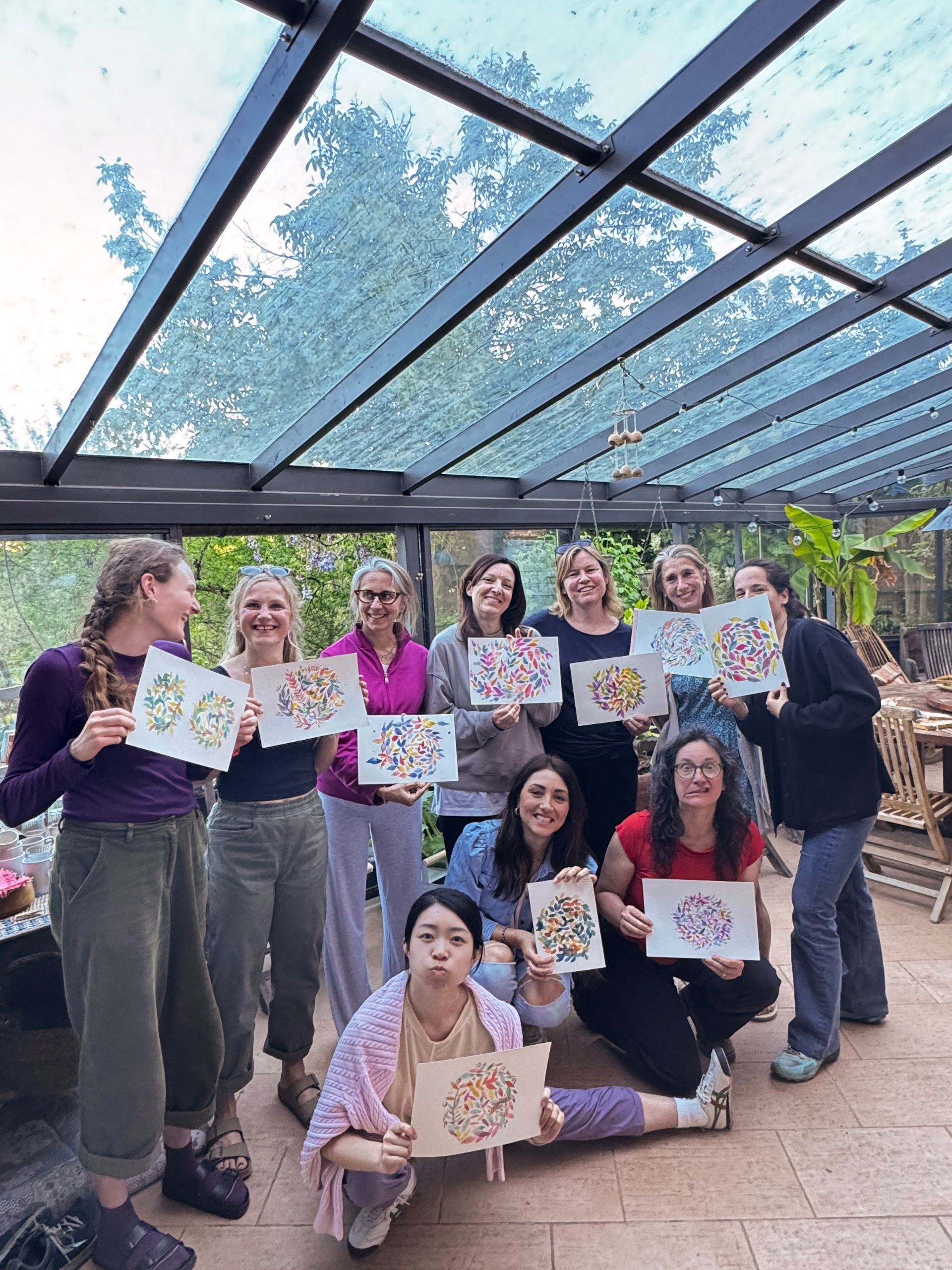 Group of women holding up colorful art in a greenhouse setting. Many are smiling, posing.