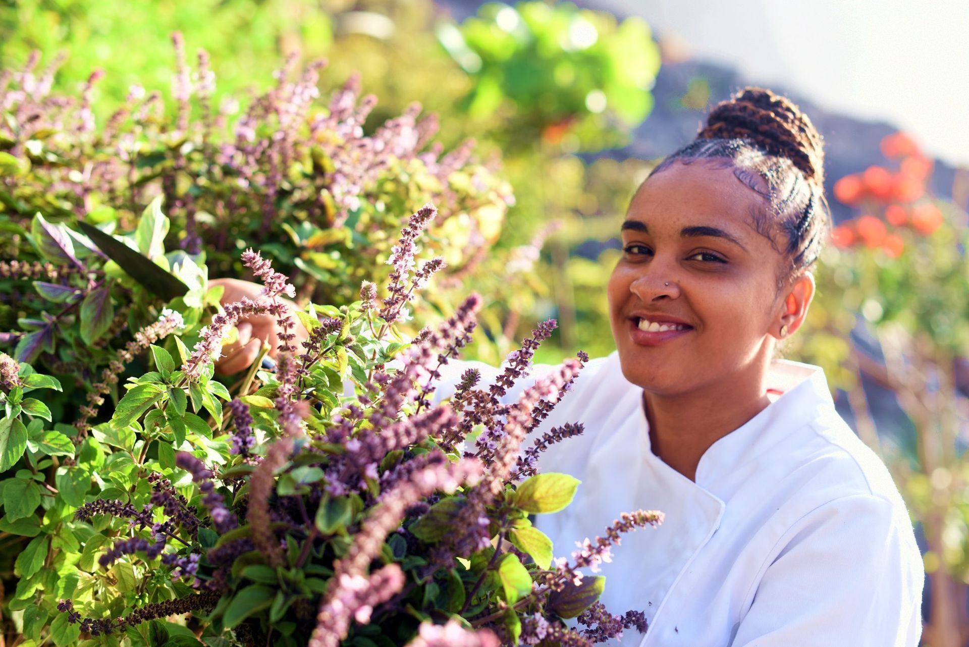 Woman in white chef coat smiles next to herb plants with purple flowers. Outdoor setting.