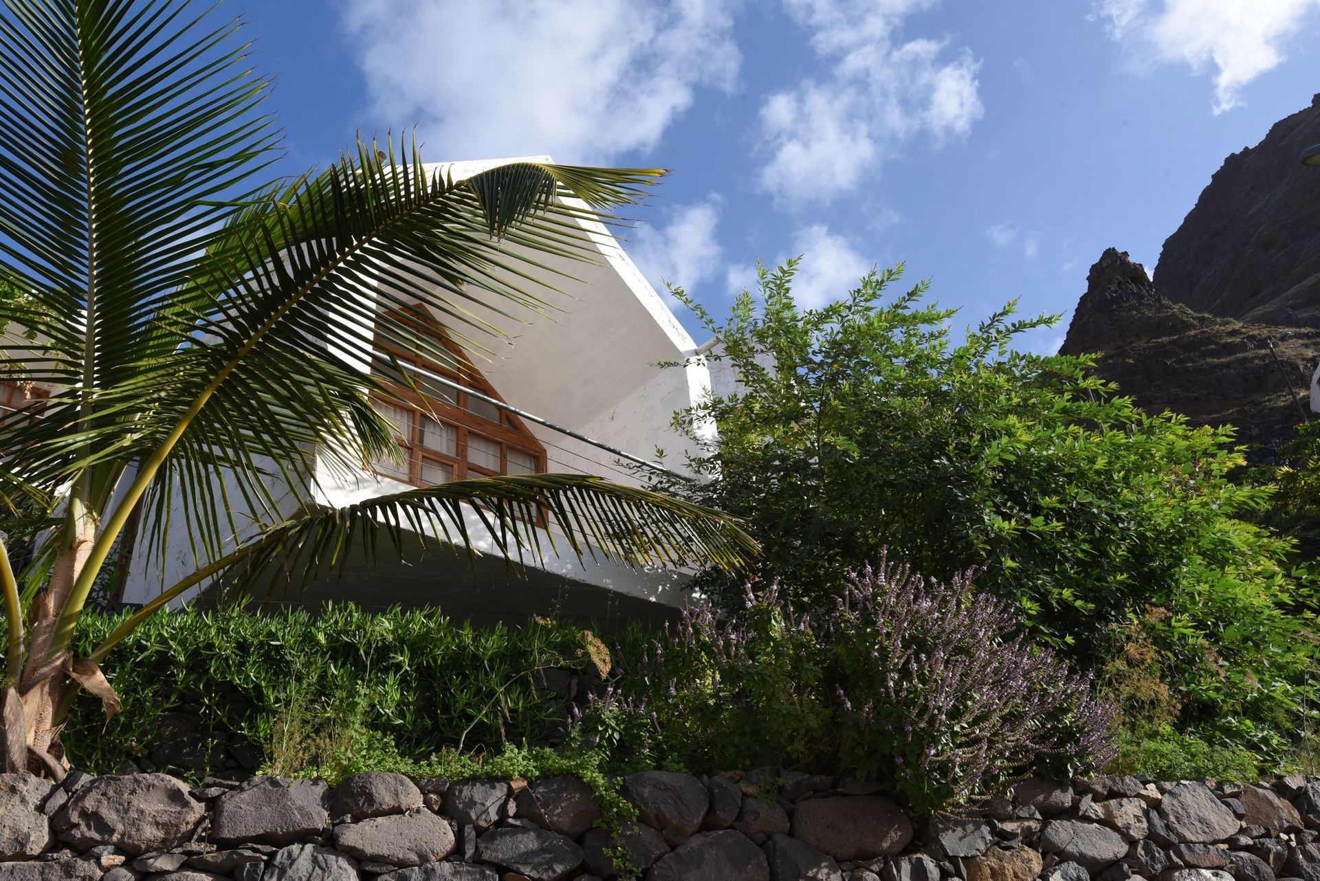 White building nestled in greenery, with stone wall in foreground, against a mountainous backdrop under a blue sky.