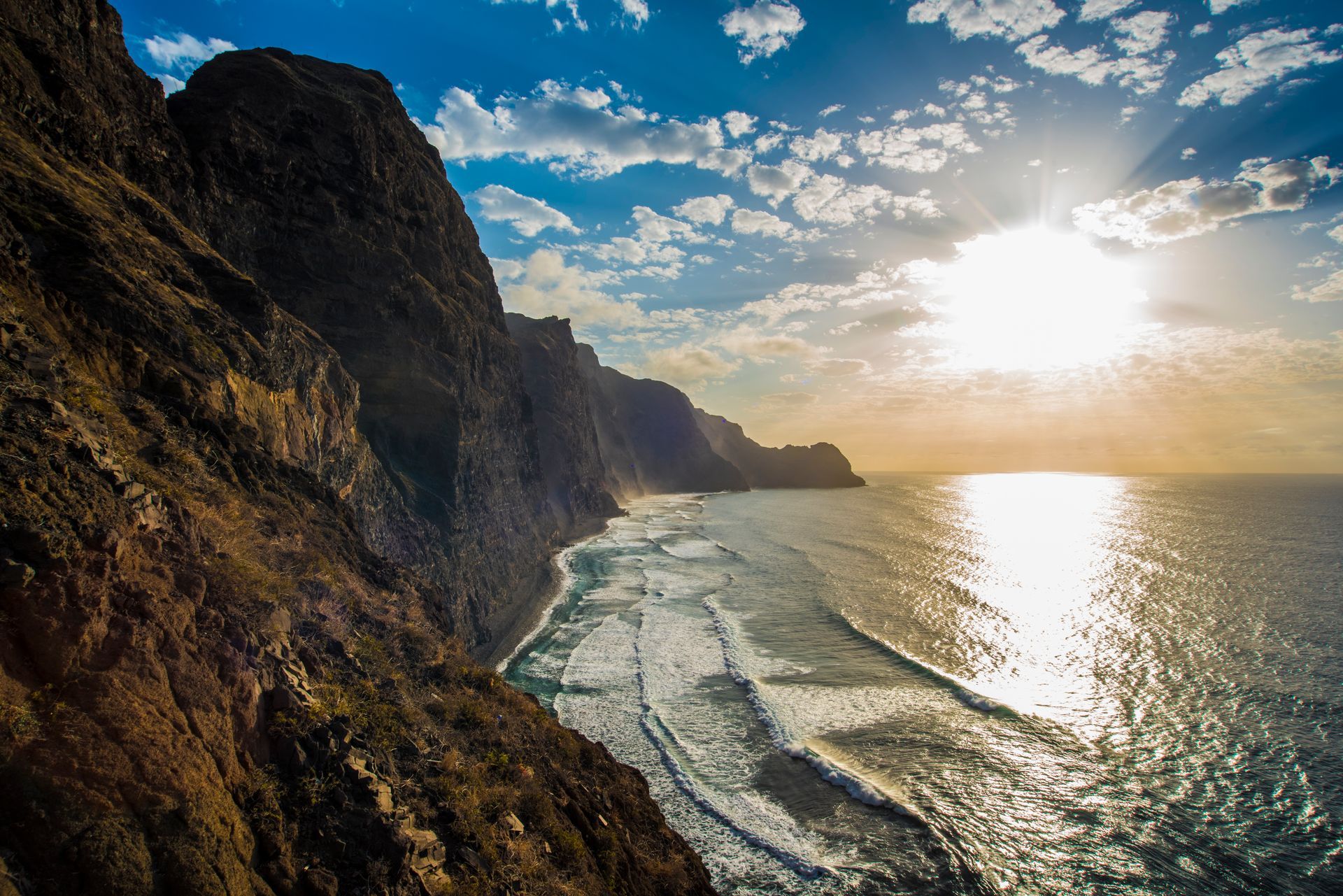 Coastal cliffs with waves crashing, bright sun shining over the ocean.