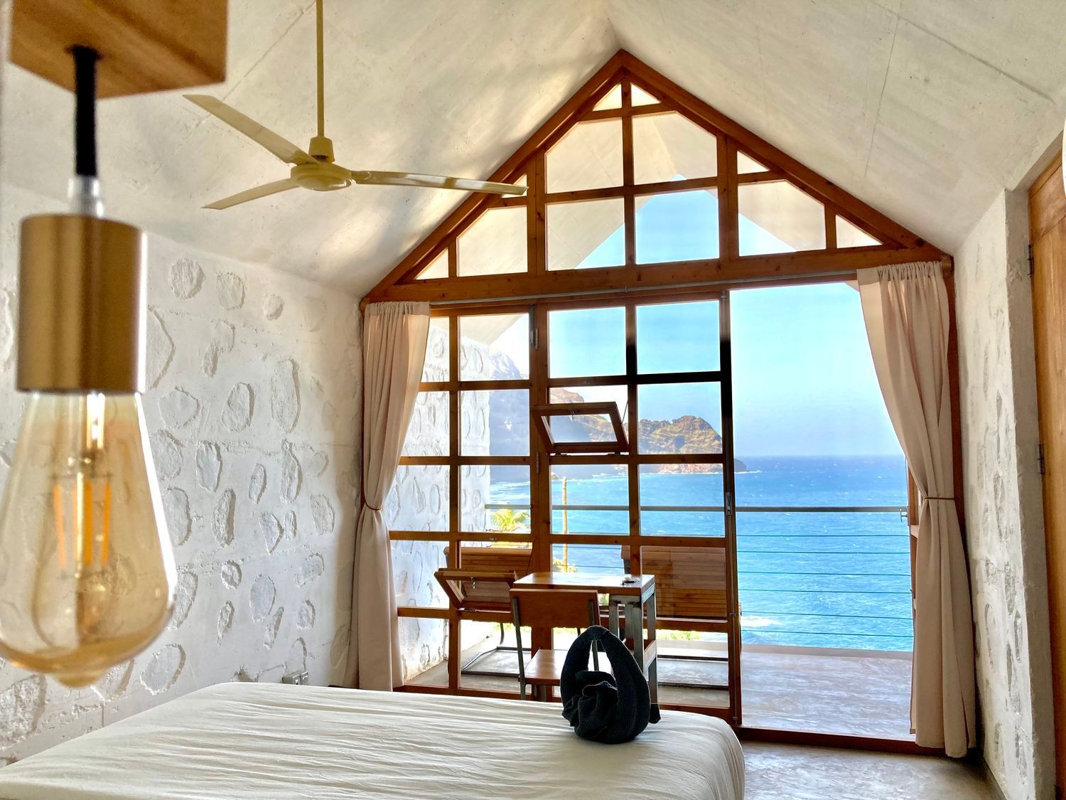 Bedroom with ocean view; textured white walls, large window, hanging light, bed, and ceiling fan.