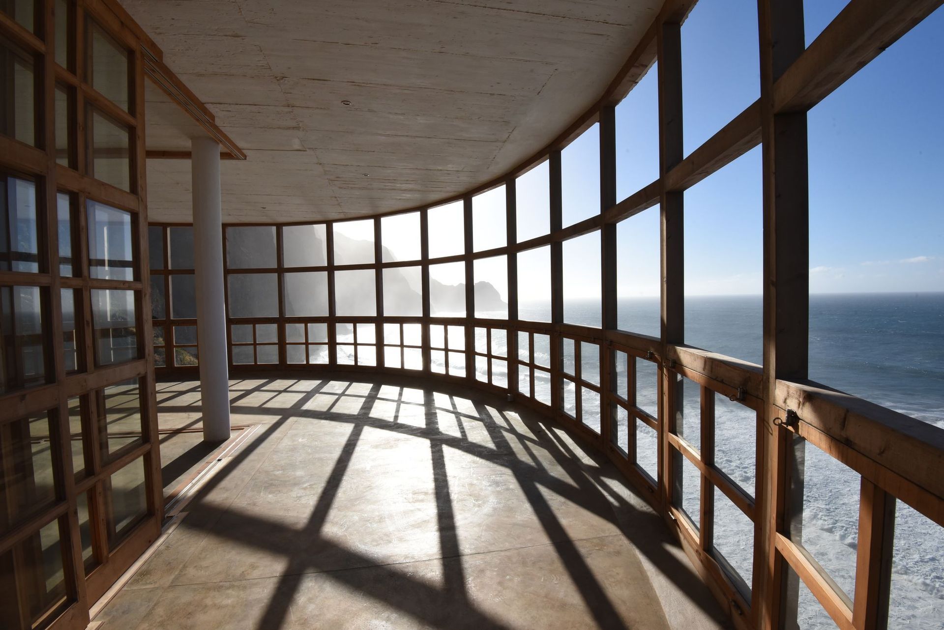 Curved balcony with wooden frames and large windows, overlooking ocean on a sunny day.