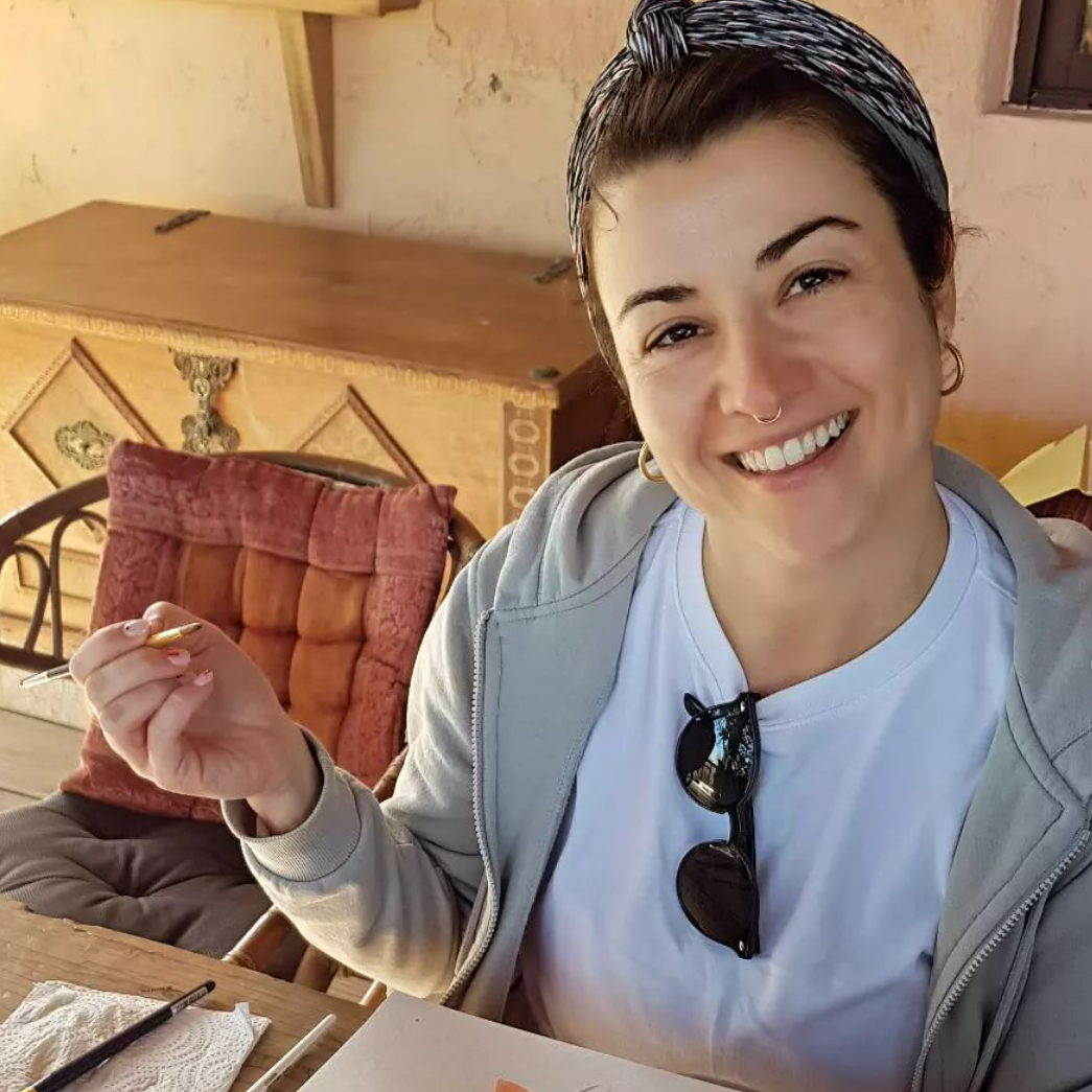 Woman with a headband smiles, holding a pen. She's seated outside, wearing a hoodie, with sunglasses on her shirt.