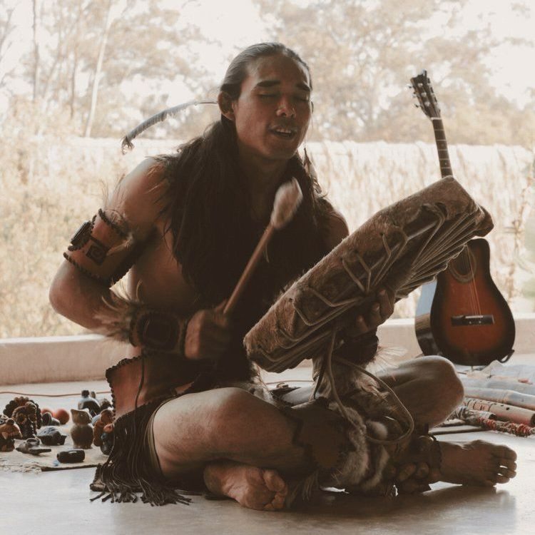 Man playing a drum while seated, in front of a window. Guitar in the background, surrounded by artifacts.