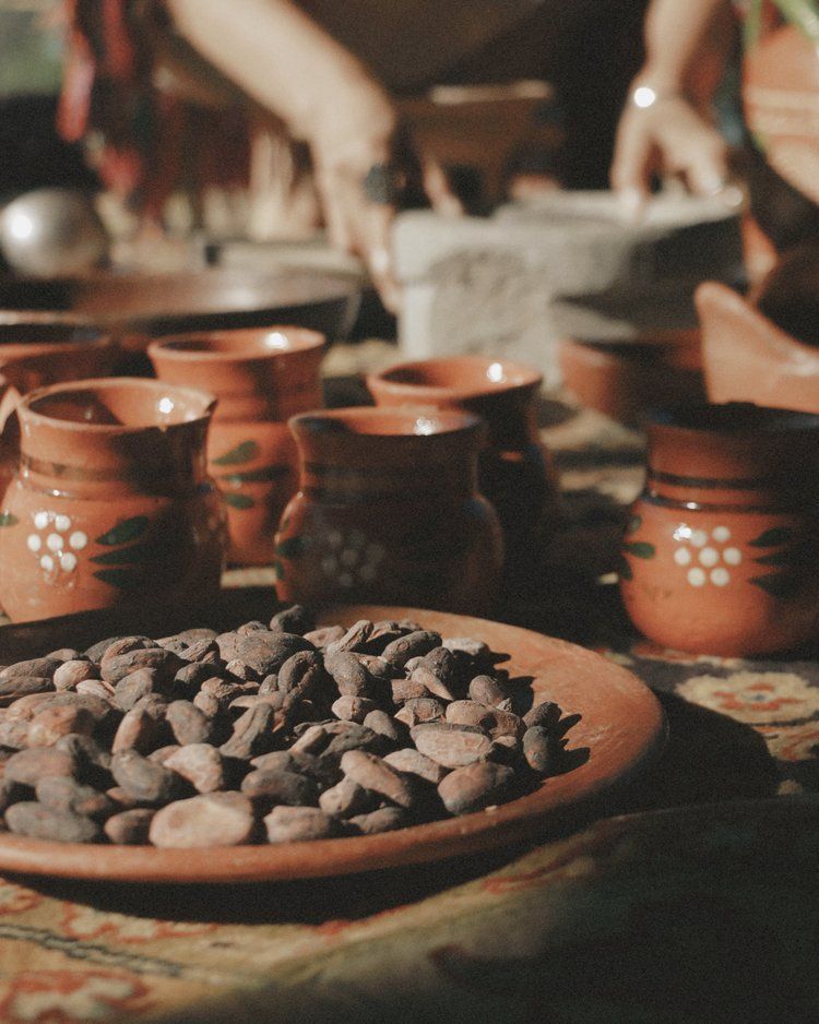 Plate of cocoa beans in front of terracotta mugs; person grinds ingredients in background.