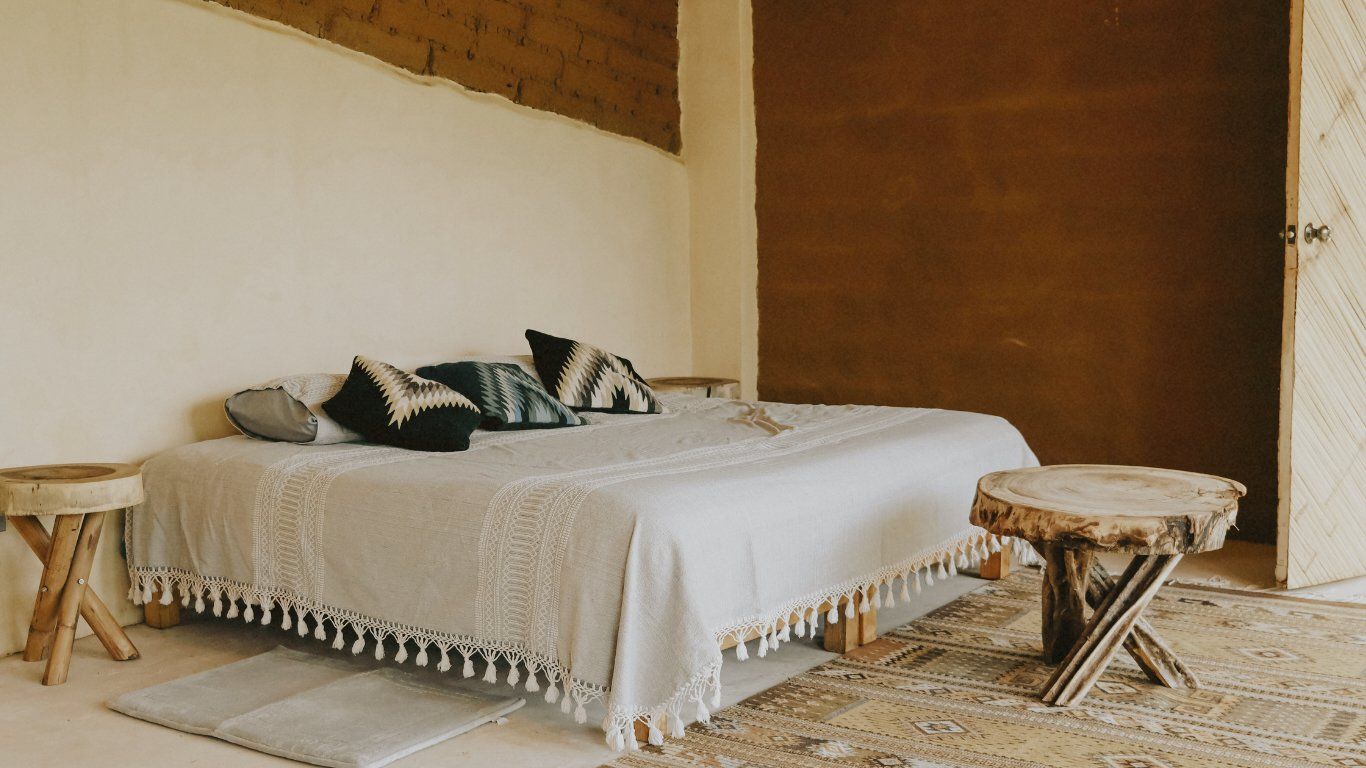 Bedroom with bed, neutral colors. Natural wood stools and woven blanket.