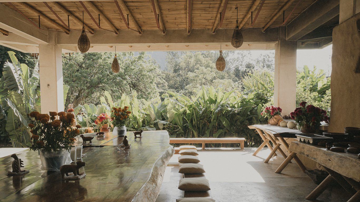Covered patio with a long wooden table set with vases. Lush greenery visible in the background.