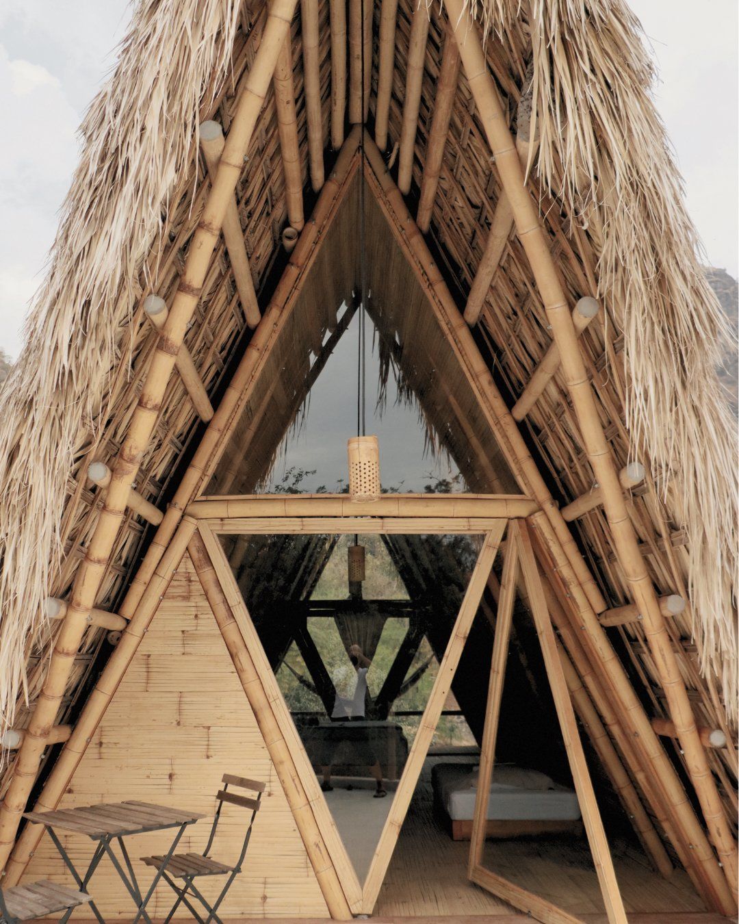 Bamboo A-frame cabin with thatched roof; small table and chairs in front. Interior visible with bed.