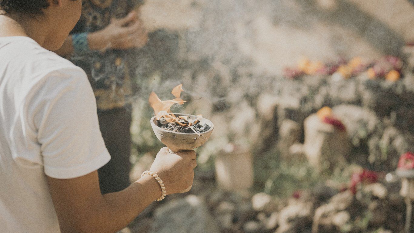 Person holding a smoking bowl of incense outdoors, near flowers and rocks.